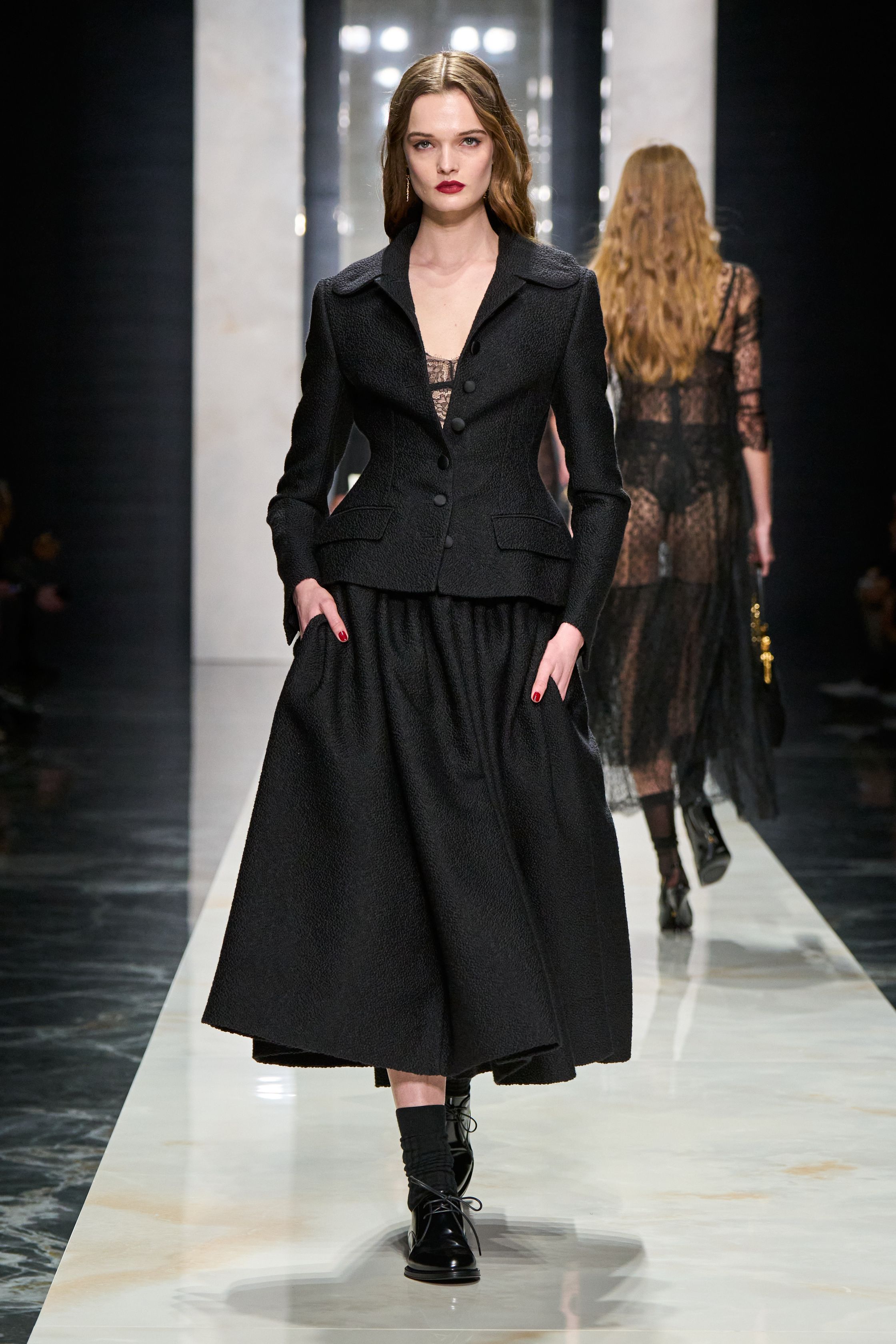 Dolce & Gabbana在本季回归品牌标志性的性感意大利美学，通过黑色蕾丝、修身西装与透视礼服塑造极具戏剧张力的女性形象。系列以黑色为核心色调，结合紧身胸衣结构、透明蕾丝裙与宽肩西装，形成刚柔并济的视觉语言。皮草外套与复古帽饰则增添奢华氛围，使整体造型在性感与优雅之间保持平衡，呈现出典型的意大利电影般的华丽气质-顽石频道趋势研究